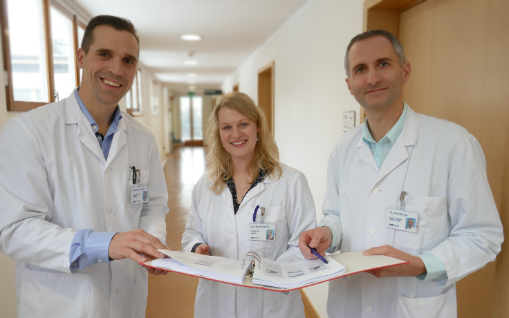 Zwei Ärzte und eine Ärztin halten einen Ordner und lächeln in die Kamera