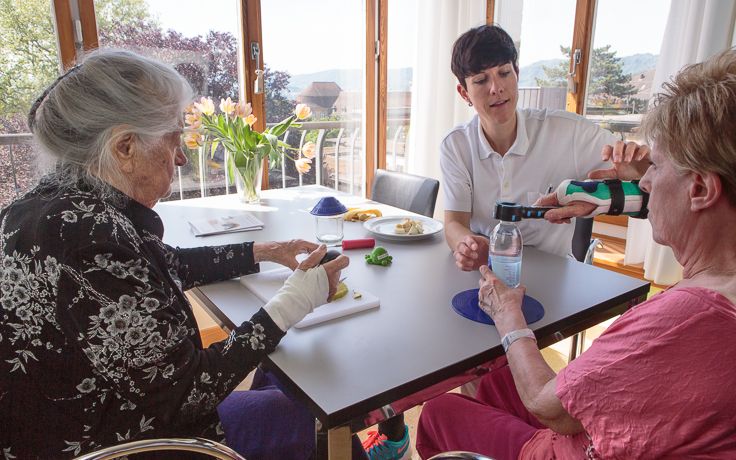 Eine Pflegerin und zwei Patientinnen sitzen an einem Tisch und trinken Wasser